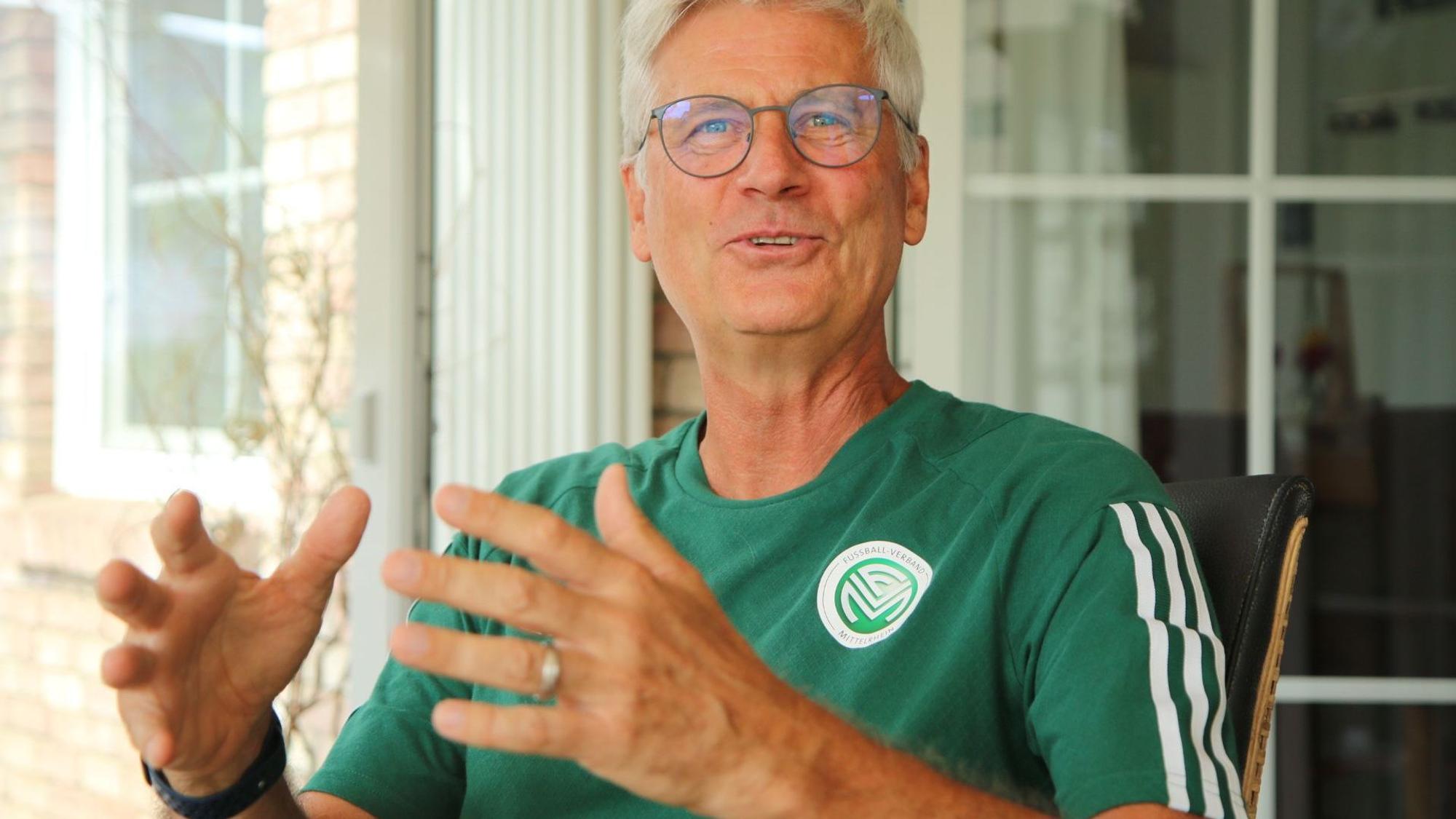 Uwe Stark sitzt gestikulierend und lachend auf einem Stuhl. Er trägt ein offizielles T-Shirt mit FVM-Logo.