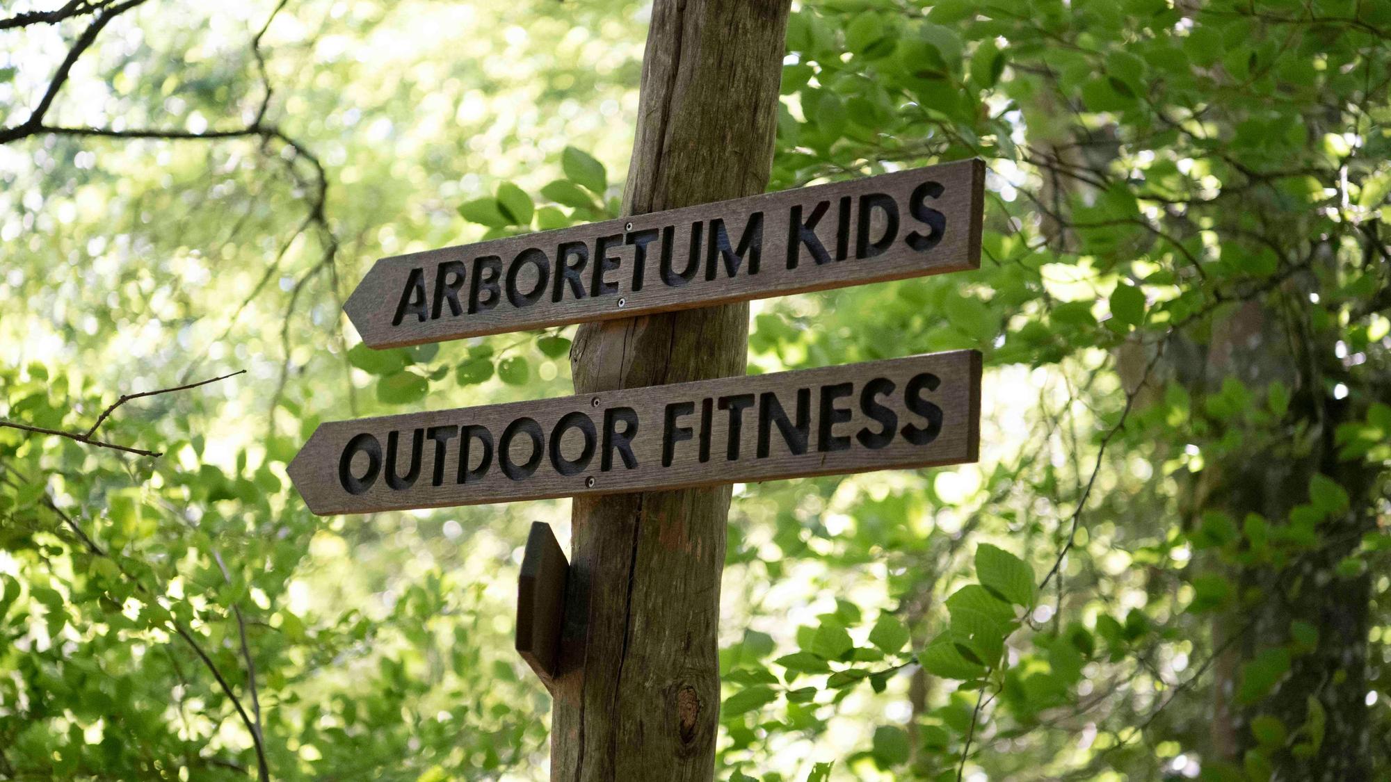 An einem Stamm in einem Wald sind zwei Hinweisschiler mit Pfeilen, auf denen „Arboretum Kids“ und „Outdoor Fitness“ eingefräst ist.