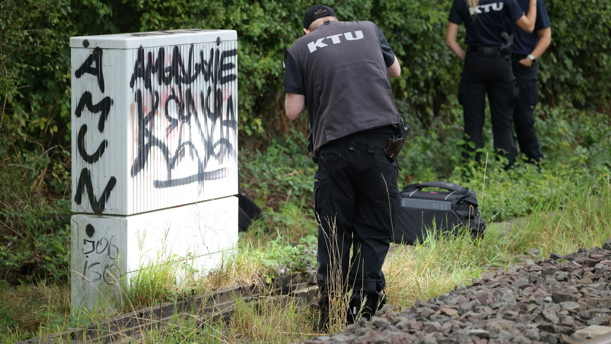 01.08.2025, Nordrhein-Westfalen, Düsseldorf: Ein Mitarbeiter der KTU (Kriminaltechnische Untersuchung) steht an «Betonwanne» am Rande der Bahngleise, in der sich verbrannte Kabel befinden. Auf die wichtige Nord-Süd-Strecke der Deutschen Bahn ist ein weiterer Brandanschlag verübt worden. Foto: Christoph Reichwein/dpa +++ dpa-Bildfunk +++
