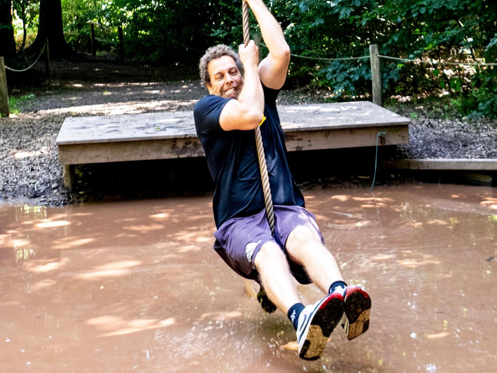 Ein Mann schwingt sich mit geschlossenen Augen und dezent verkniffenem Gesicht mit einem Tau über ein Becken mit matschigem Wasser.