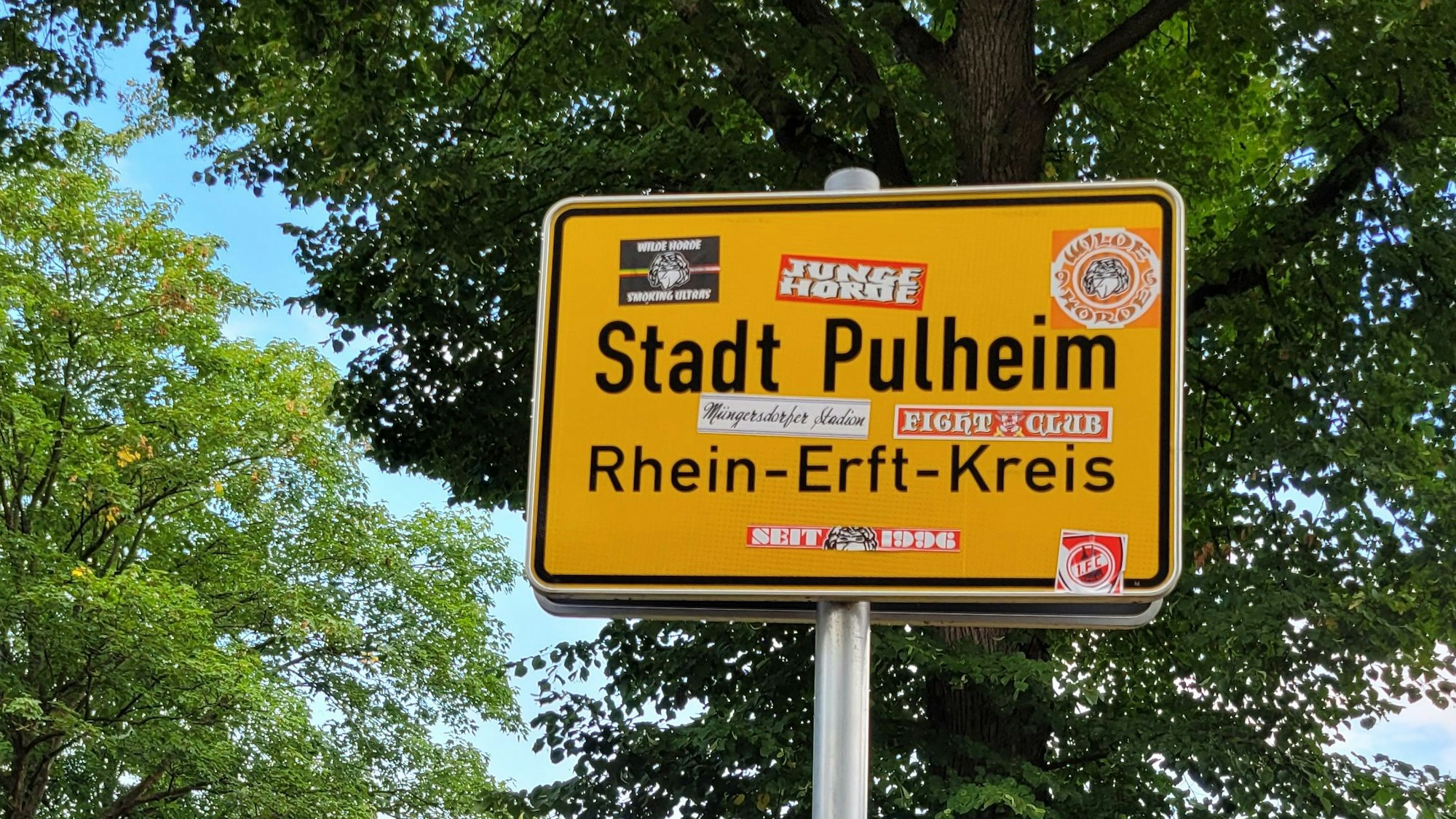 Ein Schild mit dem Schriftzug Stadt Pulheim mit vielen Aufklebern von FC-Fans.