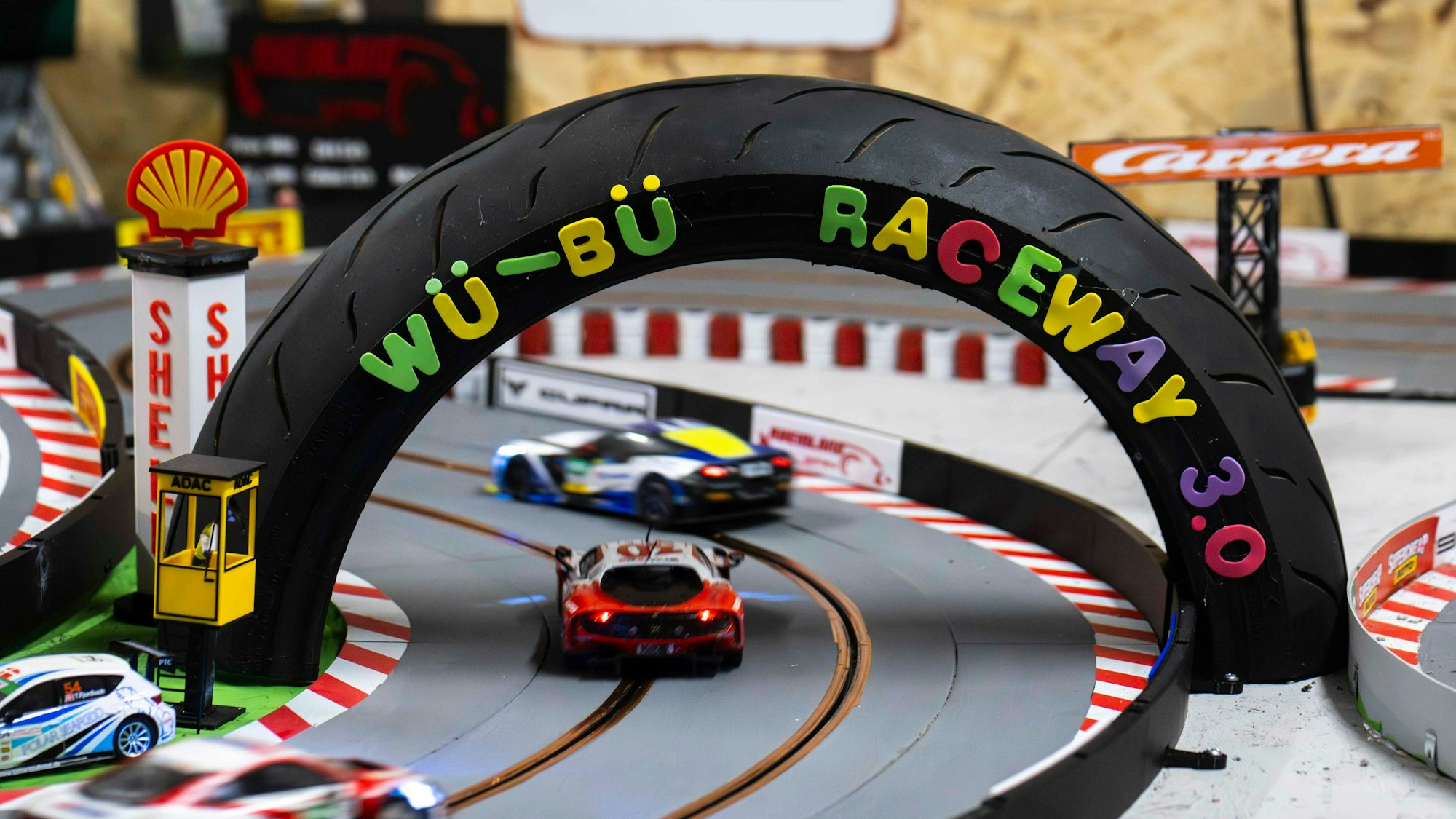Slotcars durchfahren auf ihrer Rennbahn einen in der Form eines Reifens gestalteten Bogen mit der Aufschrift „Wü-Bü Raceway 3.0“.