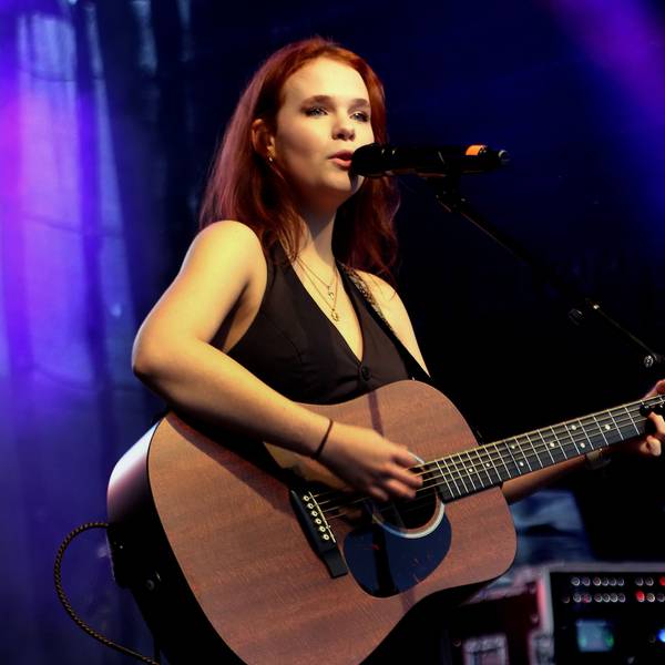 Eine junge Frau steht auf einer Bühne, sie singt und spielt Gitarre.