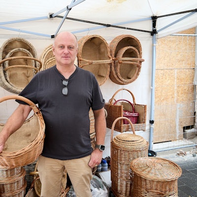 Mit einem Weidenkorb am Arm steht Korbflechter vor weiteren geflochtenen Produkten.