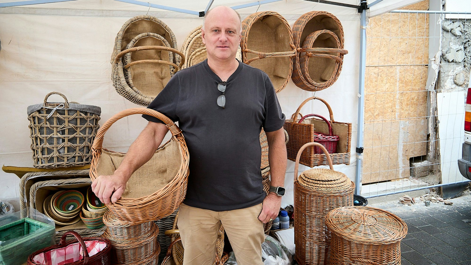 Mit einem Weidenkorb am Arm steht Korbflechter vor weiteren geflochtenen Produkten.