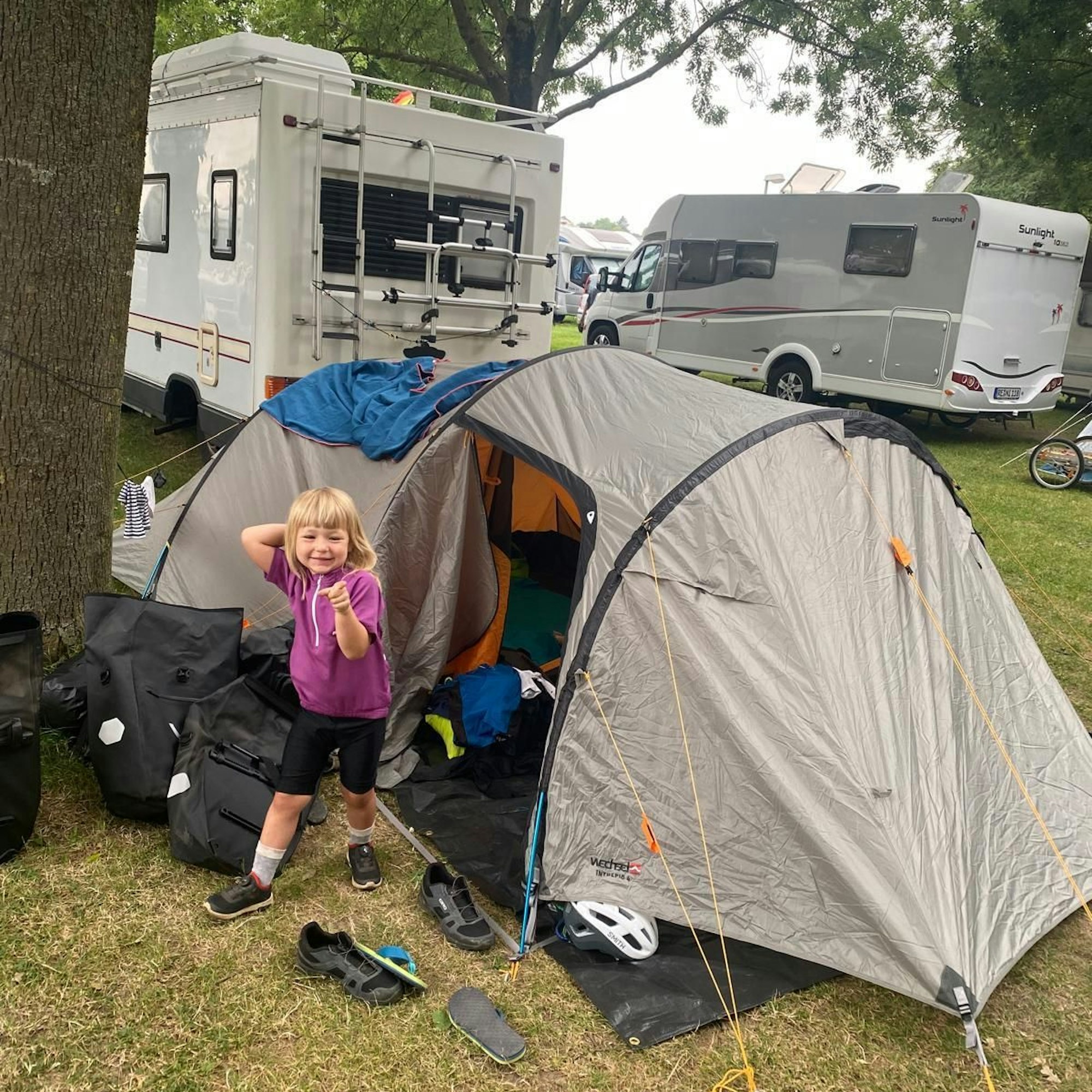 Die kleine Paula steht vor einem Zelt. Im Hintergrund sind zwei Wohnmobile zu sehen.