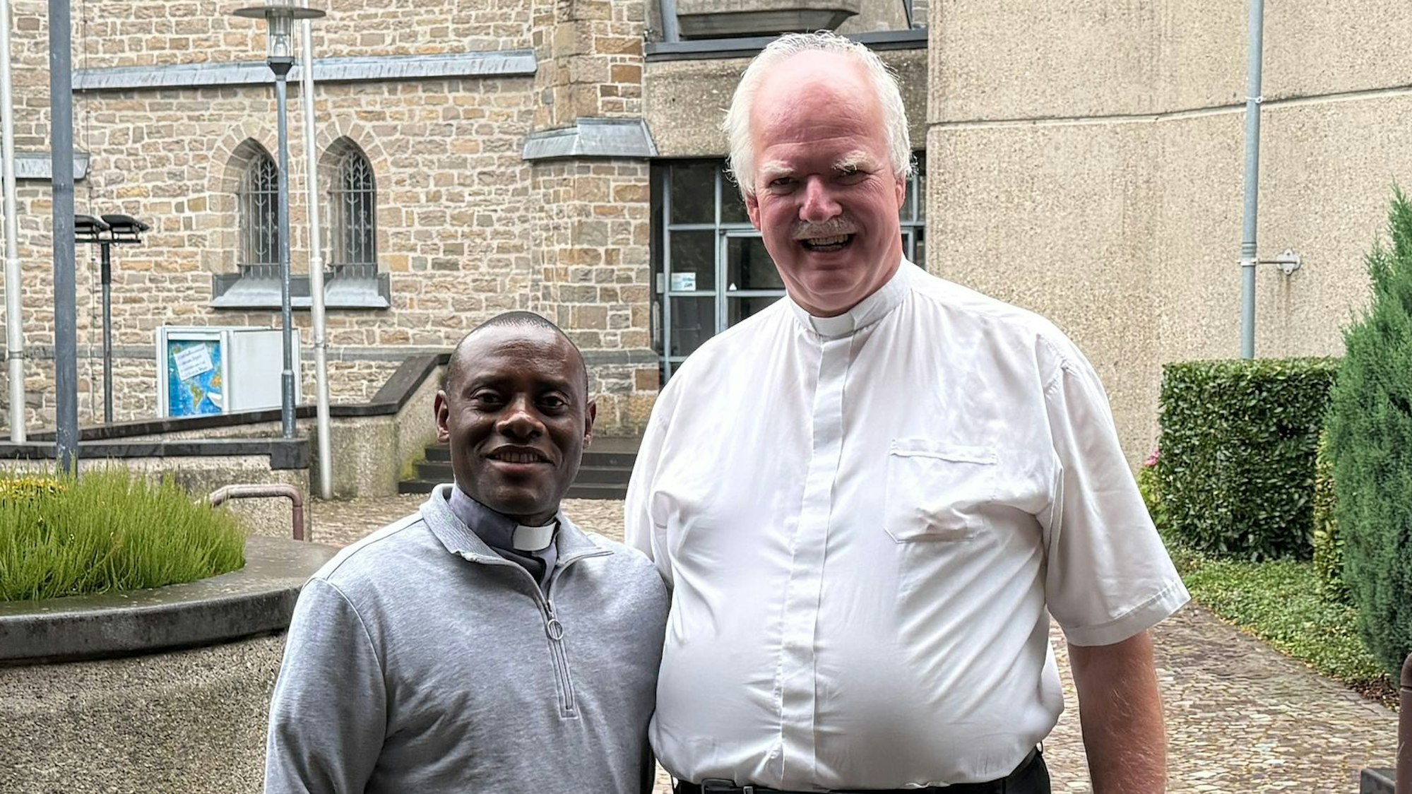 Pater Domisias Kayemba und Kreisdechant Christoph Bersch stehen vor einer Kirche nebeneinander