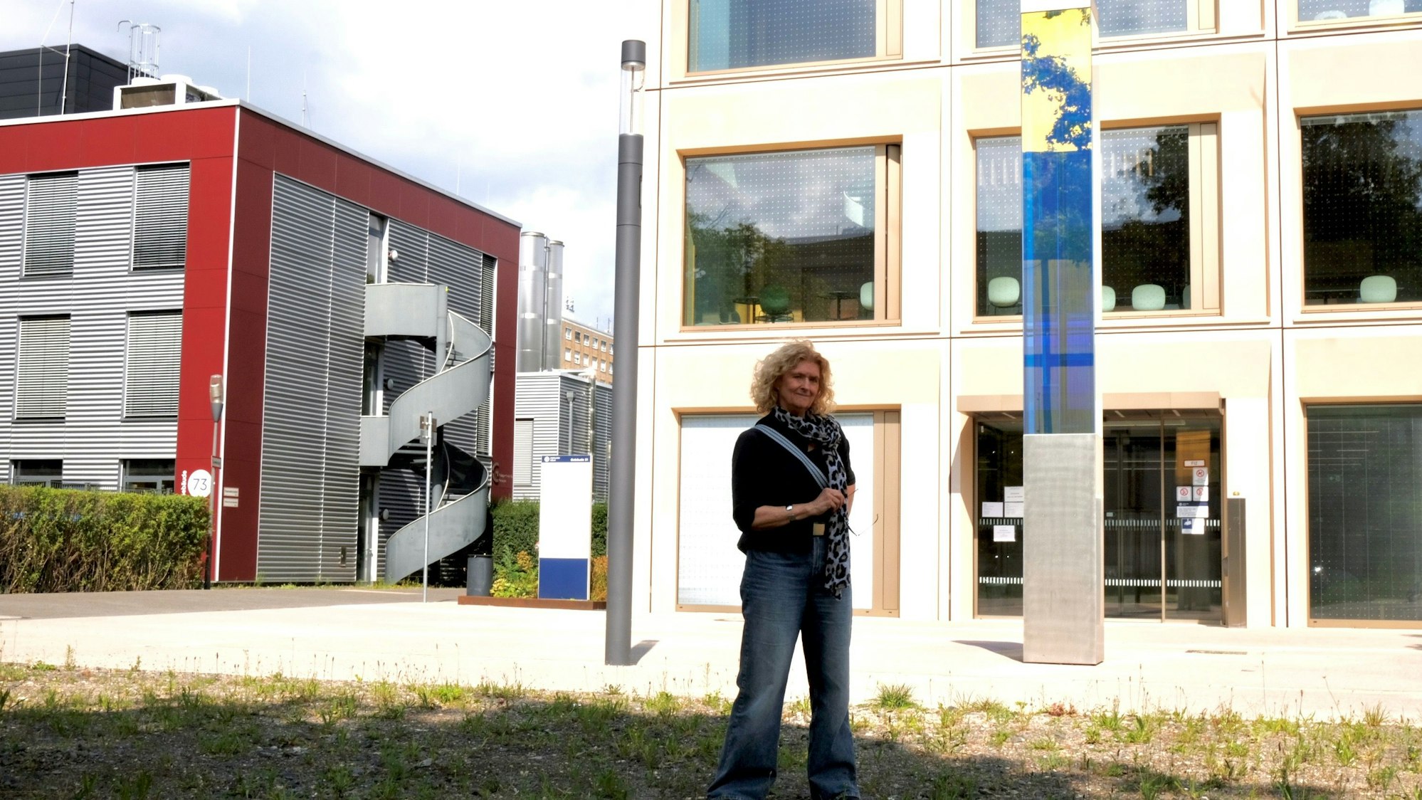 Eine Frau mit blondem Haar steht vor einem modernen Gebäude, vor dem eine Steele aus Glas und Stein steht.