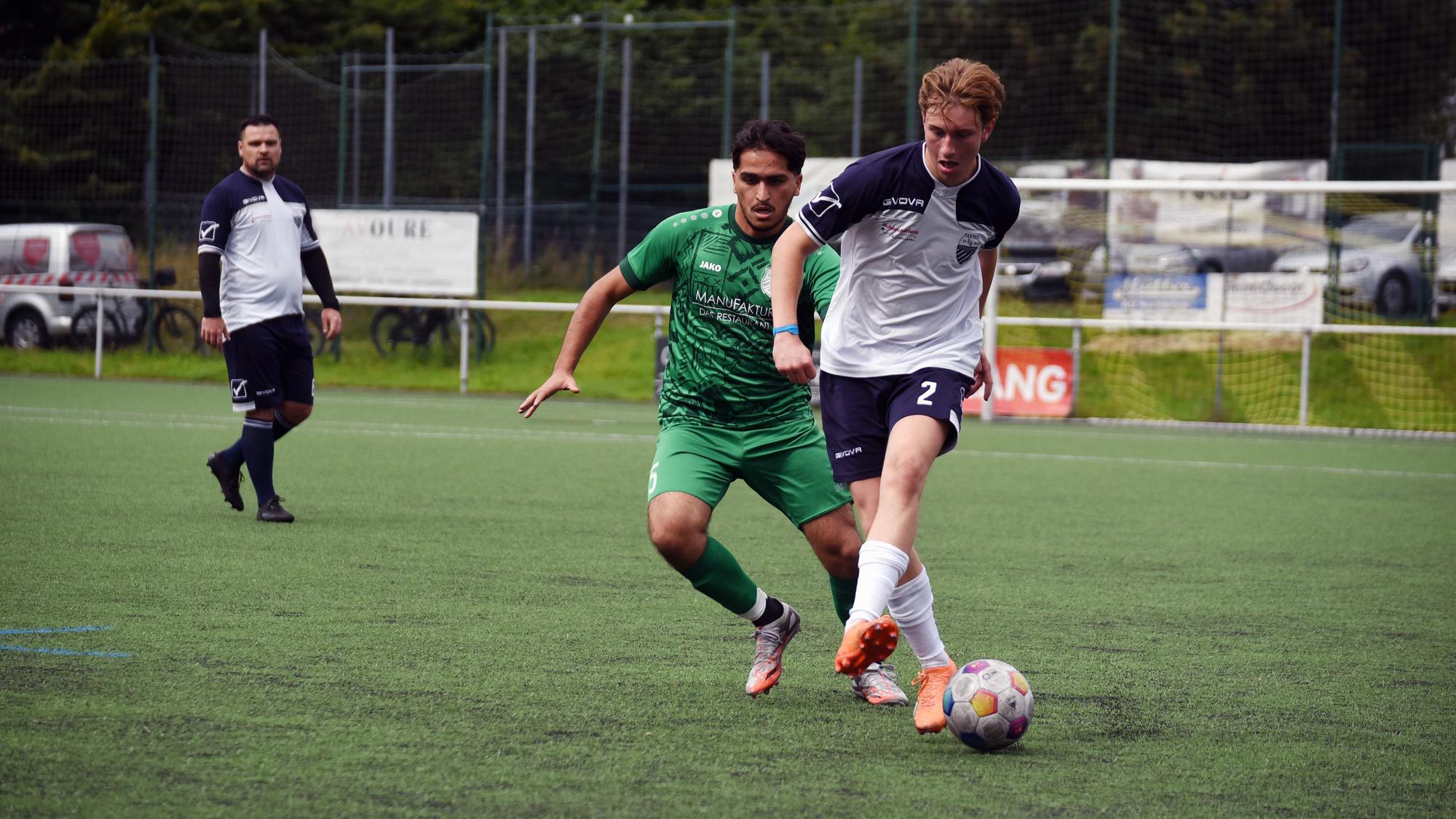 Auf einem Fußballplatz spielen zwei Mannschaften. Auf dem Bild sind zwei Spieler der jeweiligen Teams zu sehen, die um den Ball kämpfen. Im Hintergrund steht ein weiter Mitspieler.