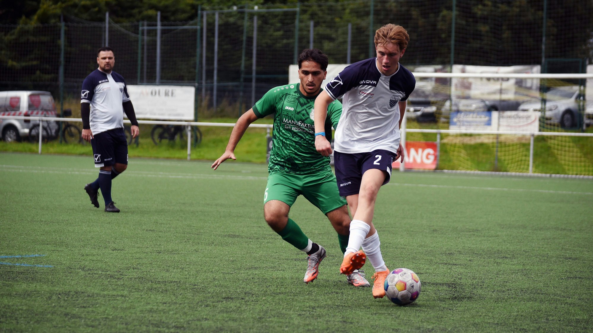 Auf einem Fußballplatz spielen zwei Mannschaften. Auf dem Bild sind zwei Spieler der jeweiligen Teams zu sehen, die um den Ball kämpfen. Im Hintergrund steht ein weiter Mitspieler.