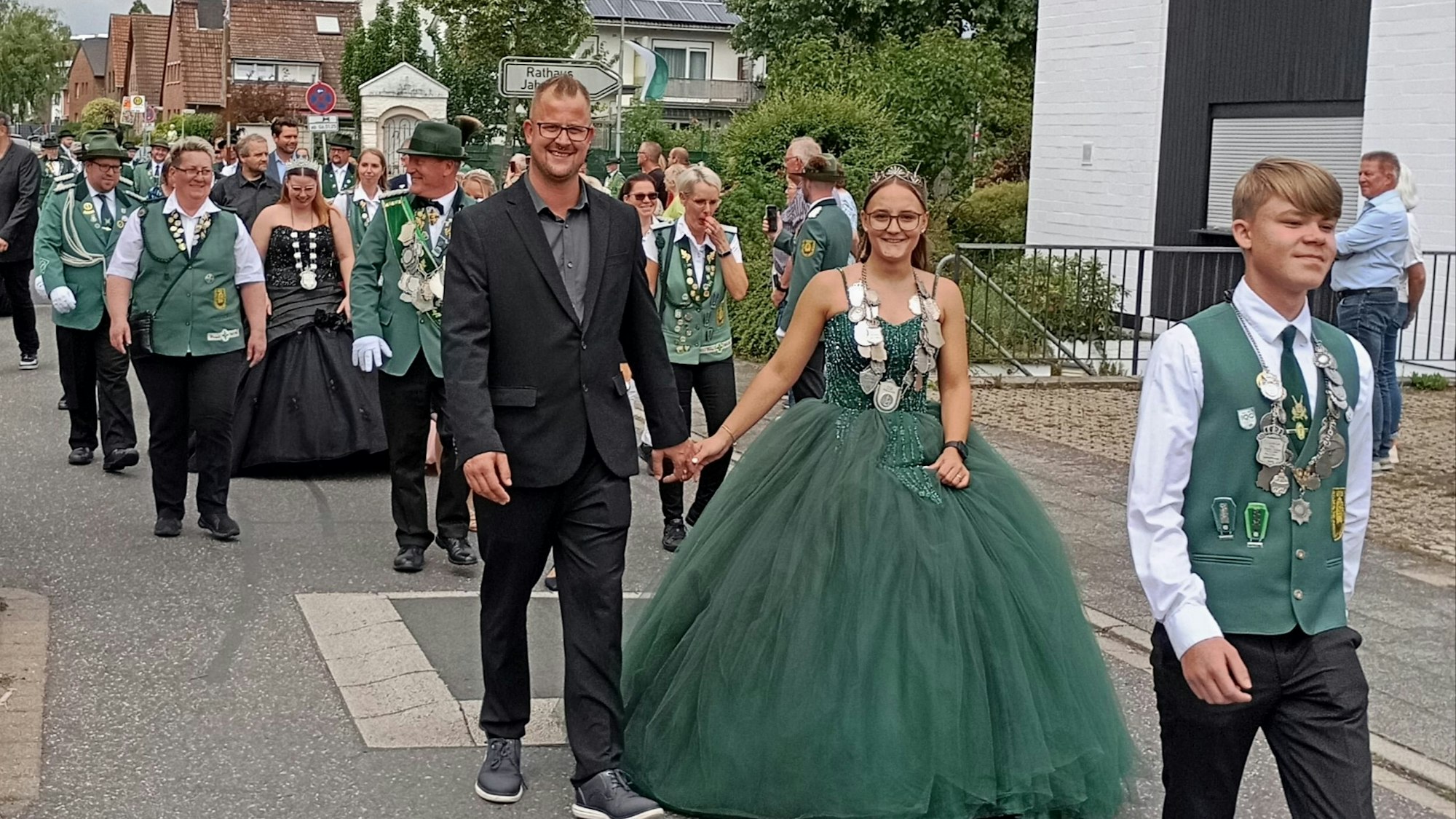 Sarah Ovenbeck trägt ein grünes Kleid, Pascal Weihrauch einen schwarzen Anzug. Die Zugteilnehmer tragen grüne traditionelle Vesten mit Ansteckern und Hüte.