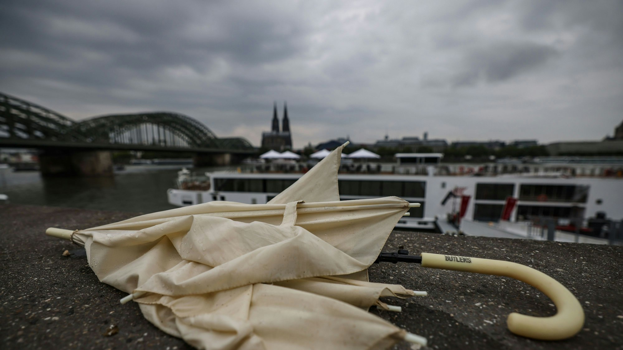 31.07.2025, Nordrhein-Westfalen, Landesweit: Ein liegen gelassener Regenschirm liegt am Rheinufer. Sonne in Nordrhein-Westfalen lässt weiter auf sich warten. Foto: Oliver Berg/dpa +++ dpa-Bildfunk +++