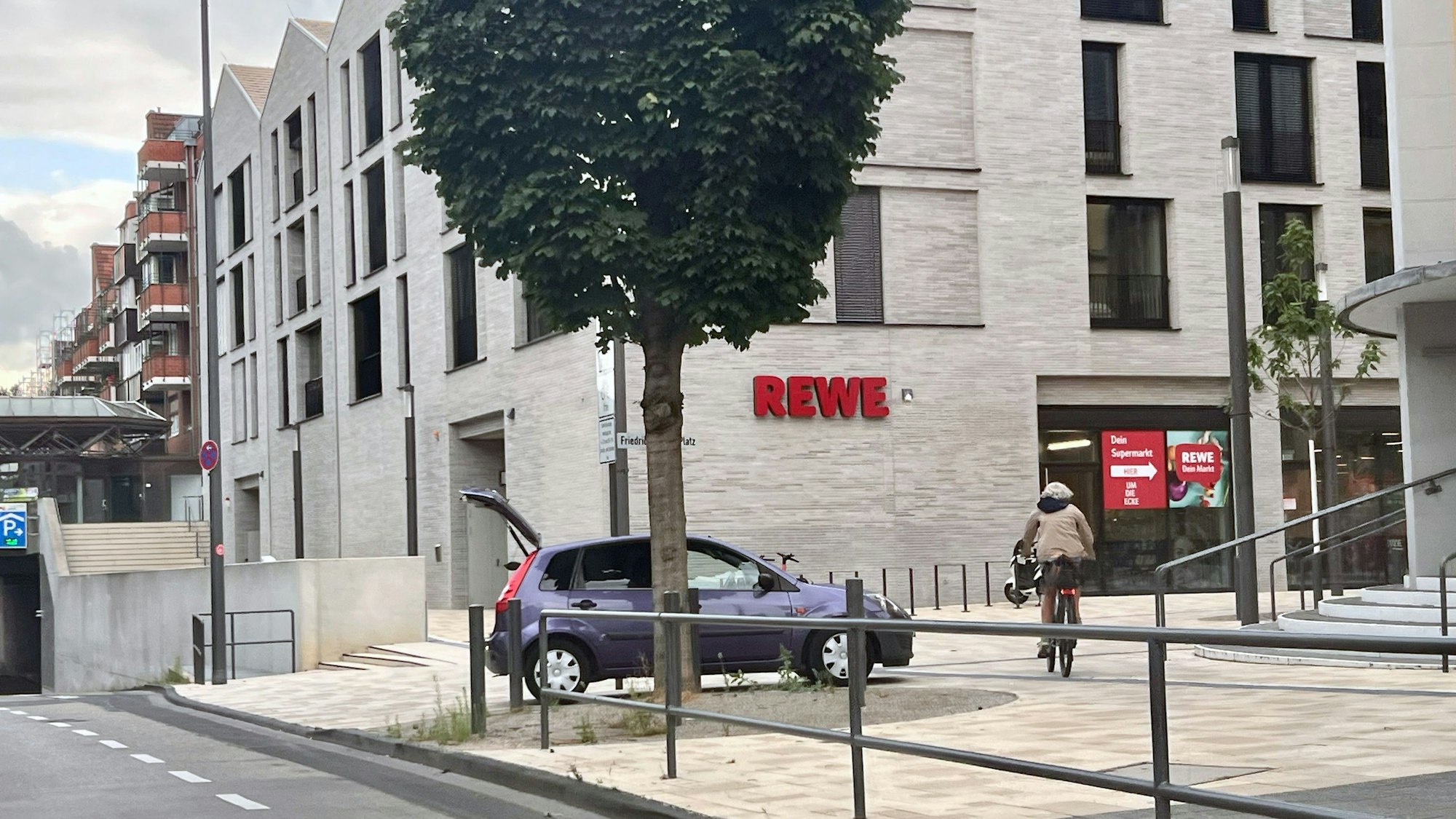 Ein neues Gebäude, auf dem REWE steht, daneben ein Auto und ein Radfahrer