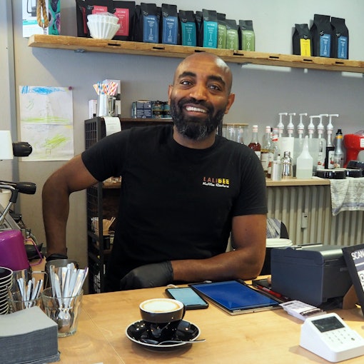 Ein Mann mit dunkler Hautfarbe steht hinter einem Café-Tresen.