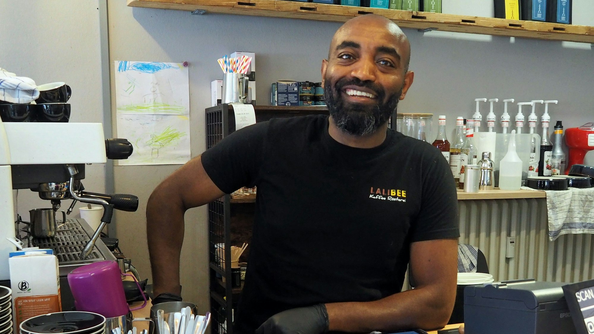 Ein Mann mit dunkler Hautfarbe steht hinter einem Café-Tresen.