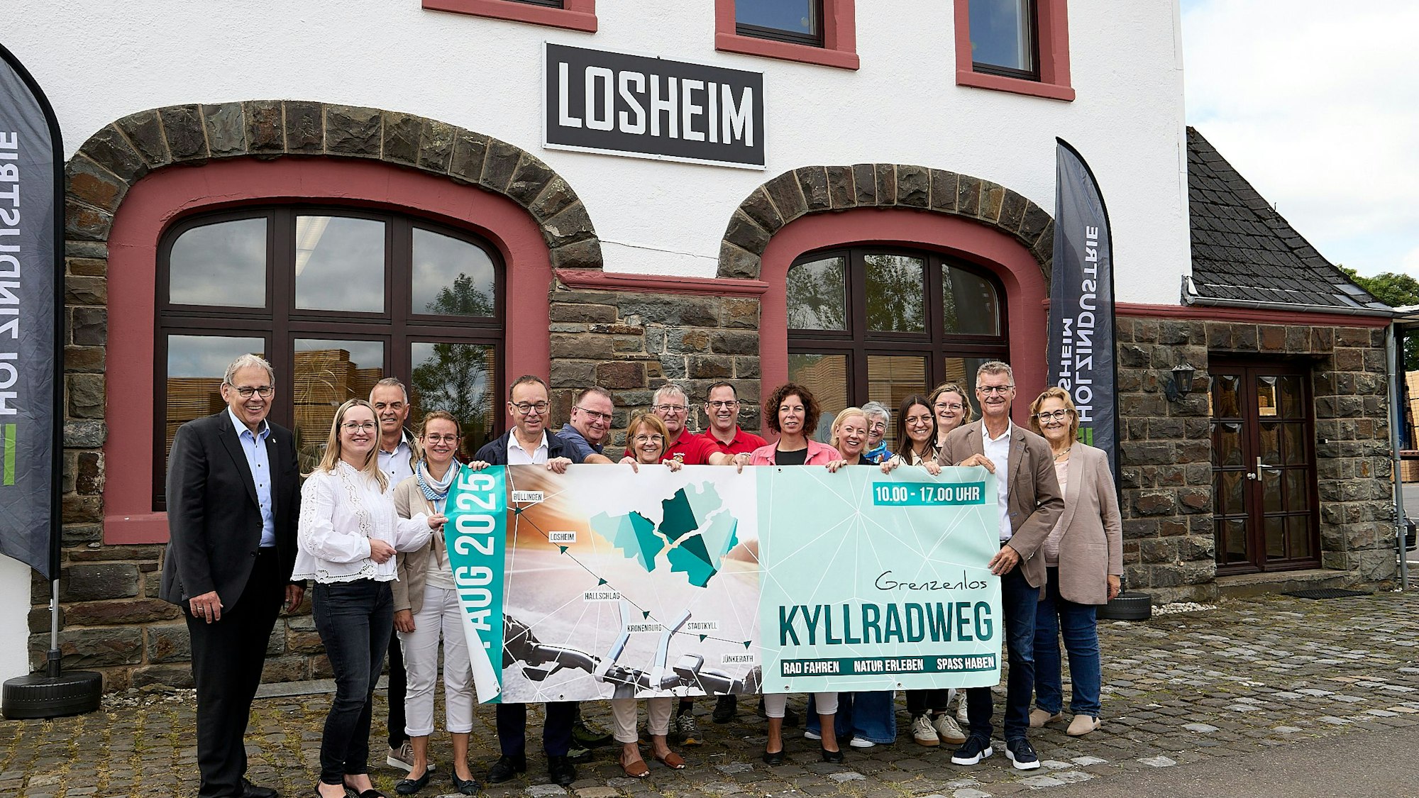 Eine Menschengruppe steht vor dem Losheimer Bahnhof und präsentiert ein Banner, auf dem „Grenzenlos Kyllradweg“ steht.