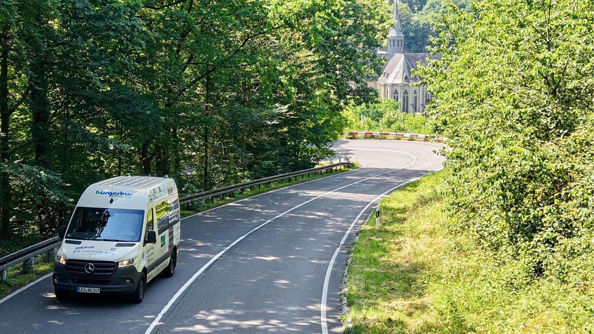 Ein kleiner Sprinter-Bus fährt durch die Serpentinen. Im Hintergrund sieht man den Altenberger Dom.