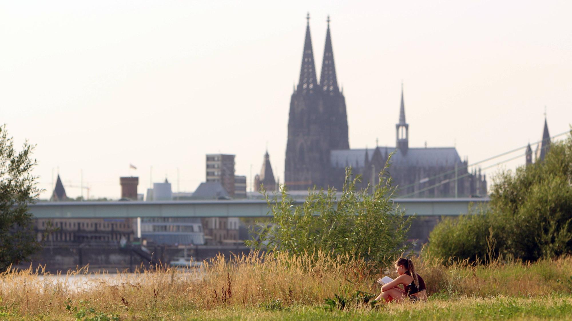 Sommerhitze in Köln am Rhein. Eine Frau entspannt sich mit einem Buch am Rheinufer in den Poller Auen vor dem Kölner Dom. (Archivfoto von 2025)