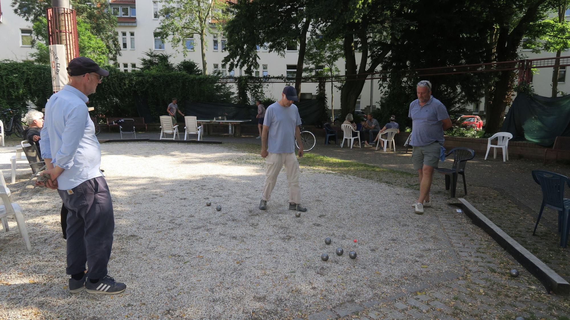 Drei Männer spielen Boule, im Hintergrund sitzen Menschen auf Plastikstühlen.