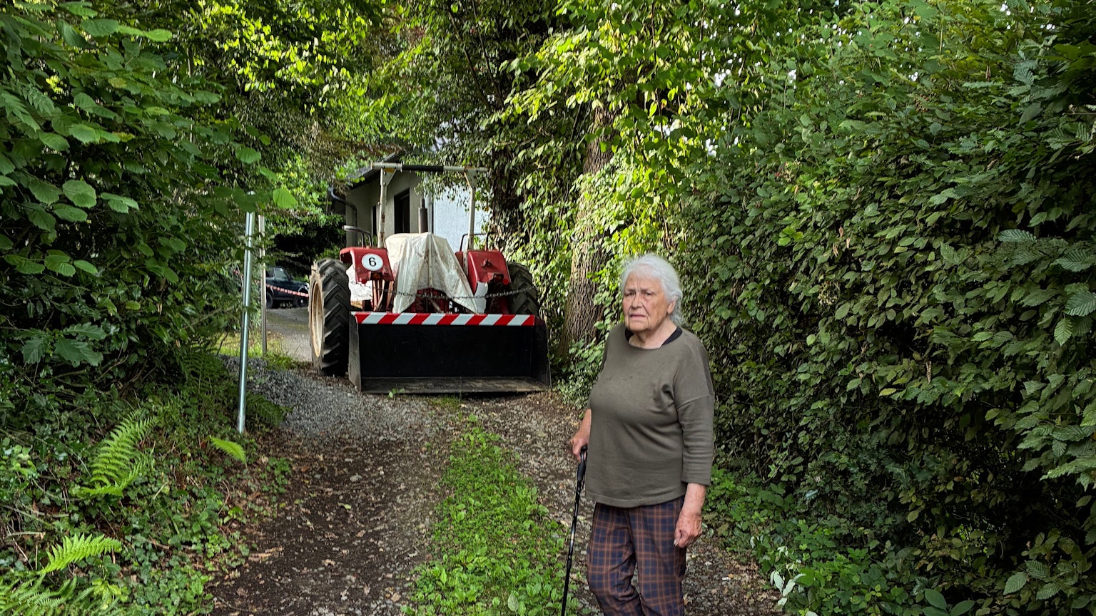 Irmintraut Fuchs steht, gestützt auf einen Stock, vor der Eisenkette und dem Trecker, die ihr den Weg zu ihrem Grundstück versperren.