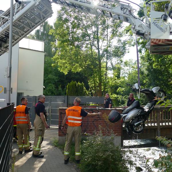 Ein Kran zieht einen Motorroller in die Höhe, eine Gruppe von Männern steht dabei, zwei von ihnen tragen orangefarbene Warnwesten mit der Aufschrift „Feuerwehr“.