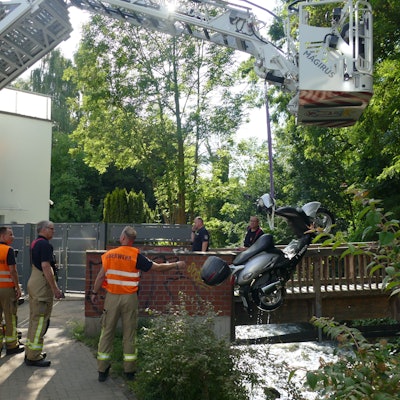 Ein Kran zieht einen Motorroller in die Höhe, eine Gruppe von Männern steht dabei, zwei von ihnen tragen orangefarbene Warnwesten mit der Aufschrift „Feuerwehr“.