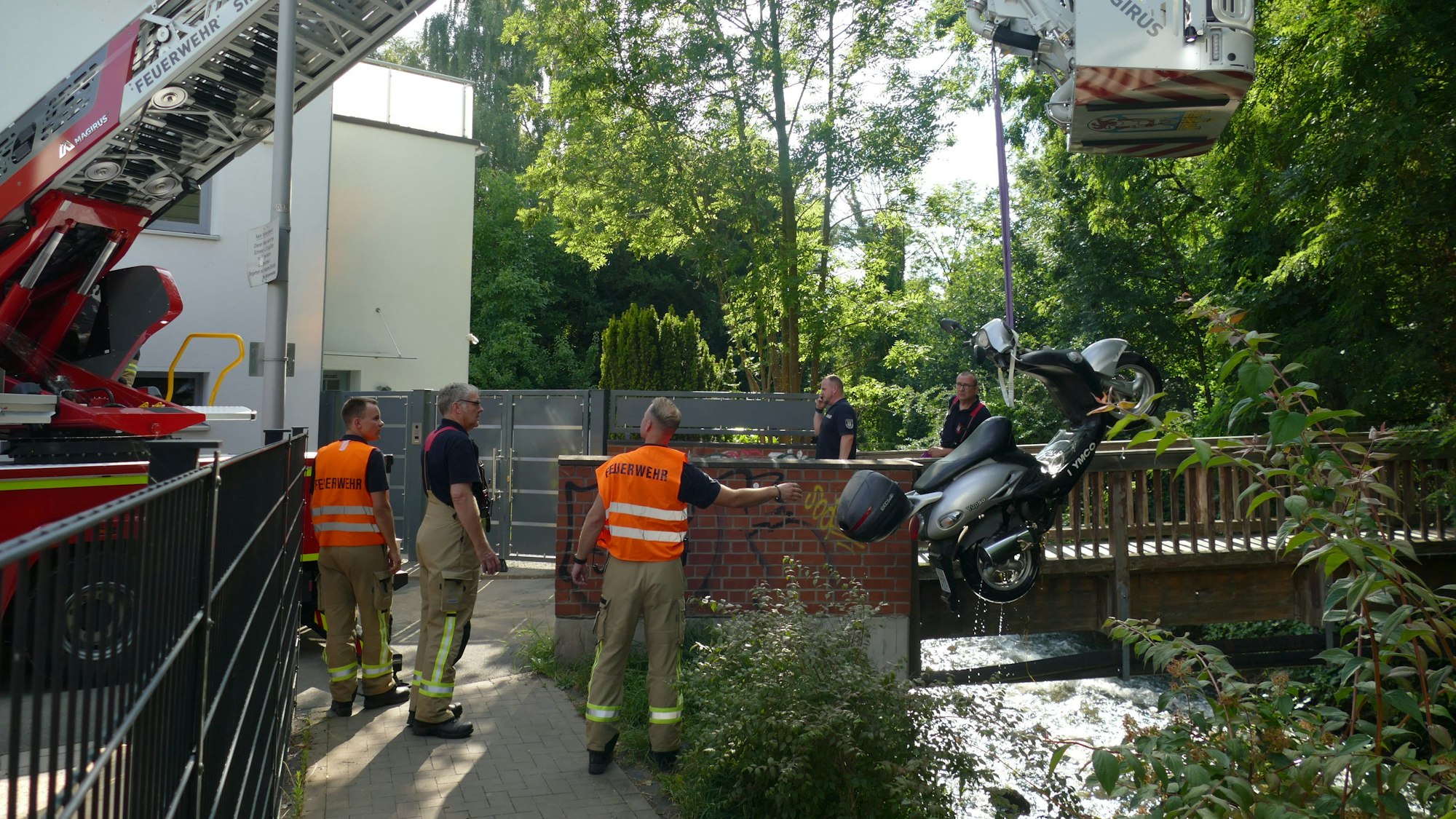 Ein Kran zieht einen Motorroller in die Höhe, eine Gruppe von Männern steht dabei, zwei von ihnen tragen orangefarbene Warnwesten mit der Aufschrift „Feuerwehr“.