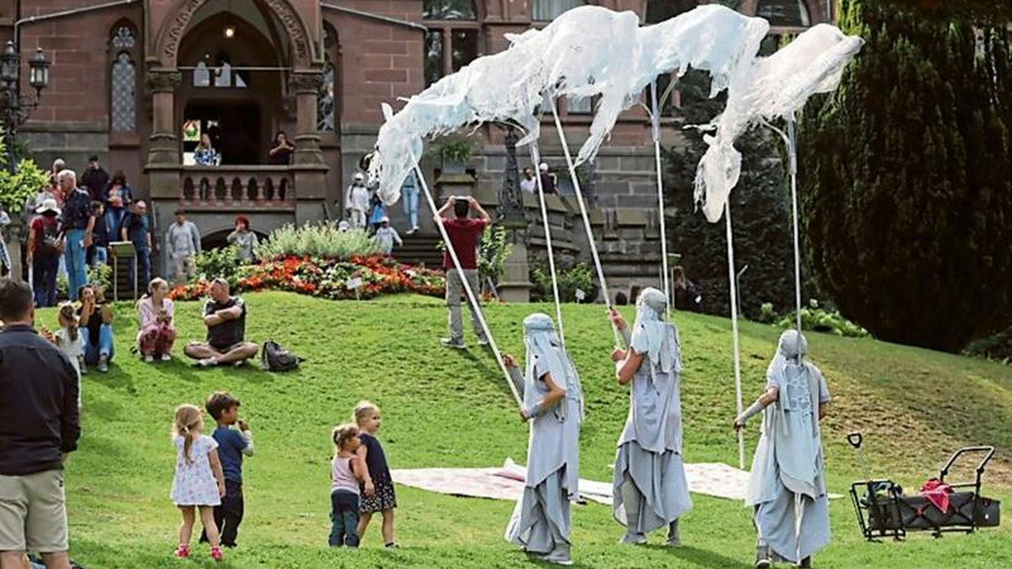 Drei verkleidete Gestalten auf einer Wiese vor einer Burg, einige Kinder schauen zu.
