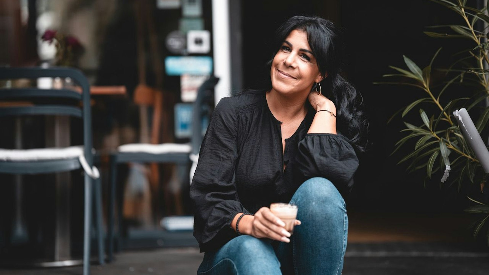 Eine Frau mit dunklem Haar im Pferdeschwanz sitzt auf den Stufen vor einer geöffneten Café-Tür.