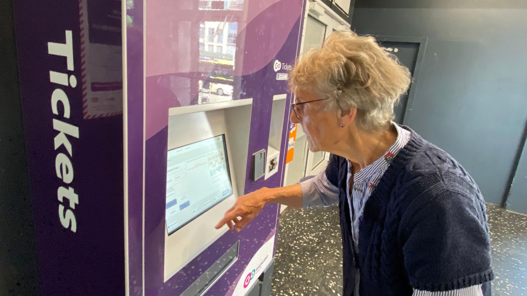 Eine ältere Dame steht an einem Ticketautomaten und schaut konzentriert auf das Display.