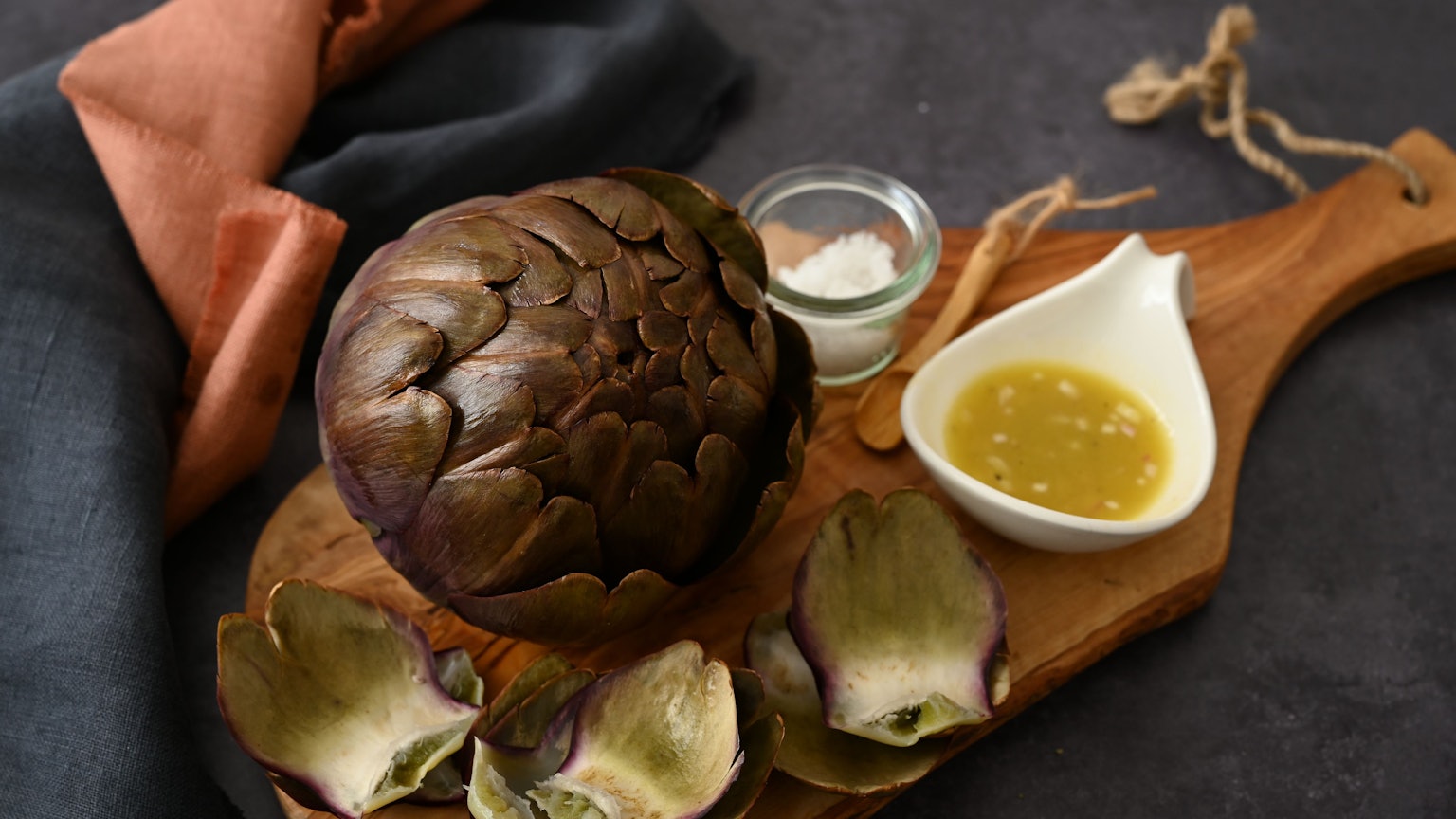 Artischocken haben jetzt Saison. Eine zubereitete Artischocke liegt auf einem Brettchen, daneben eine Schale Vinaigrette.