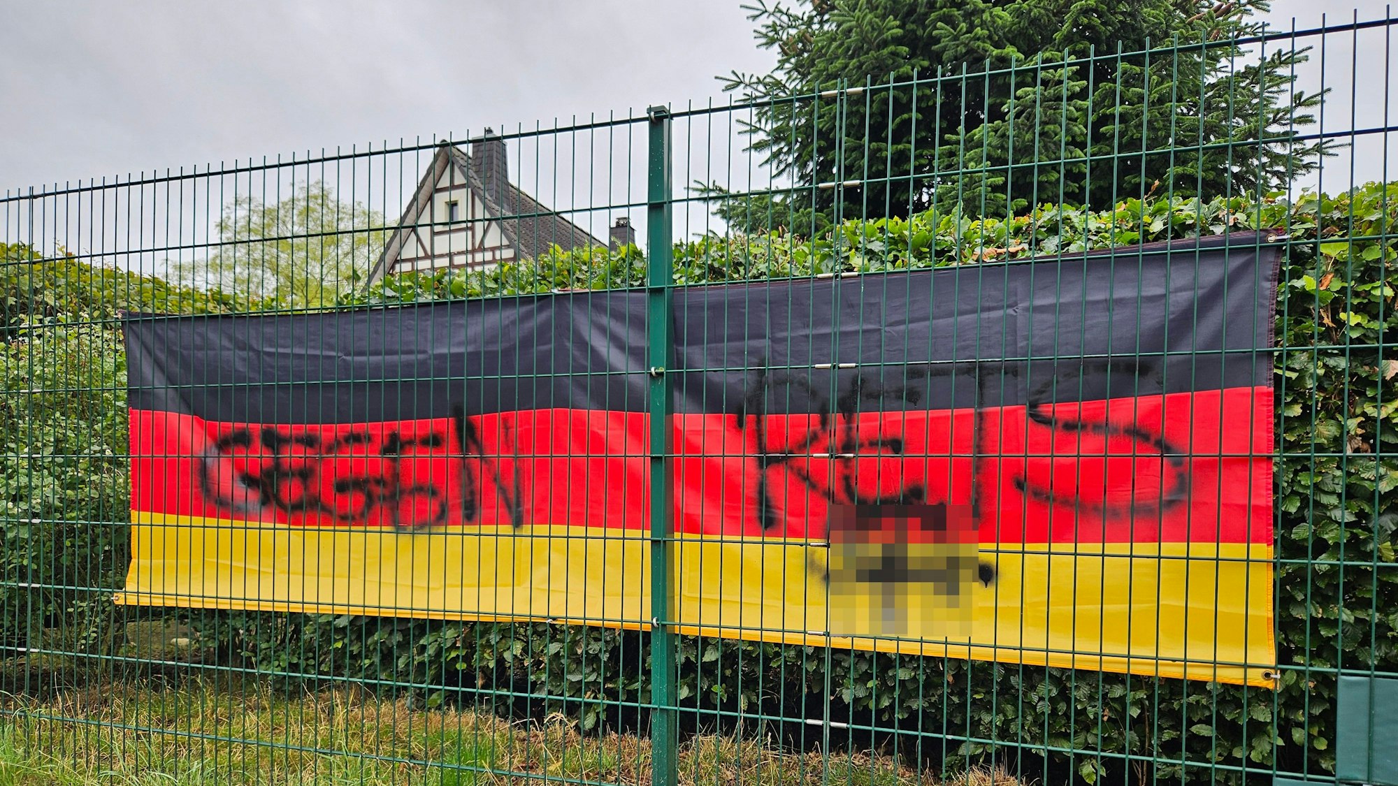 Aufnahme einer Deutschlandfahne hinter einem Zaun, auf der ein aufgesprühtes Hakenkreuz verpixelt wurde.