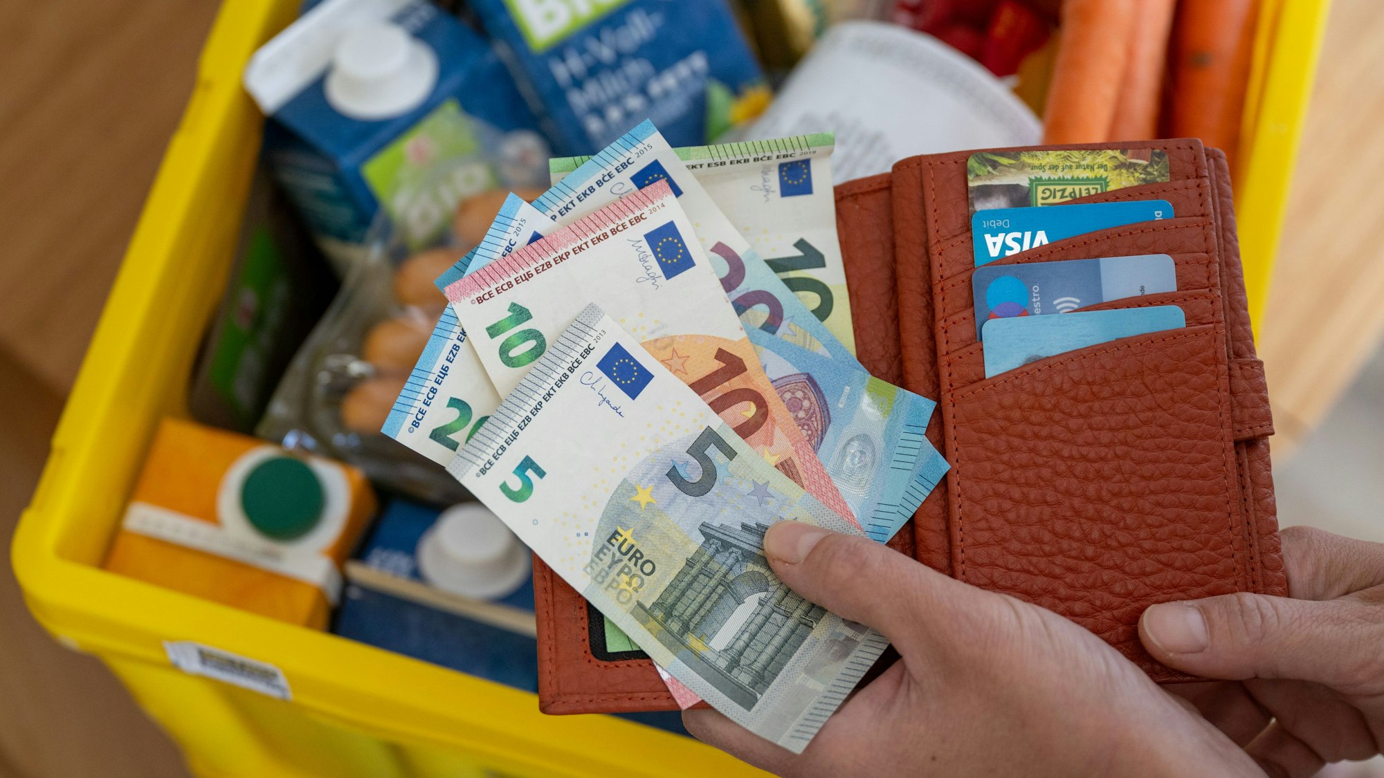 Eine Frau hält ein Portemonnaie mit Bargeld und Karten über einer Einkaufskiste mit Lebensmitteln.