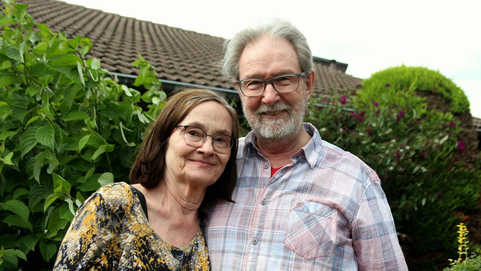 Das Paar steht vor einigen Büschen vor ihrem Haus. Werner Hehl trägt ein kariertes Hemd, Brunhilde Hehl ein gemustertes Kleid.