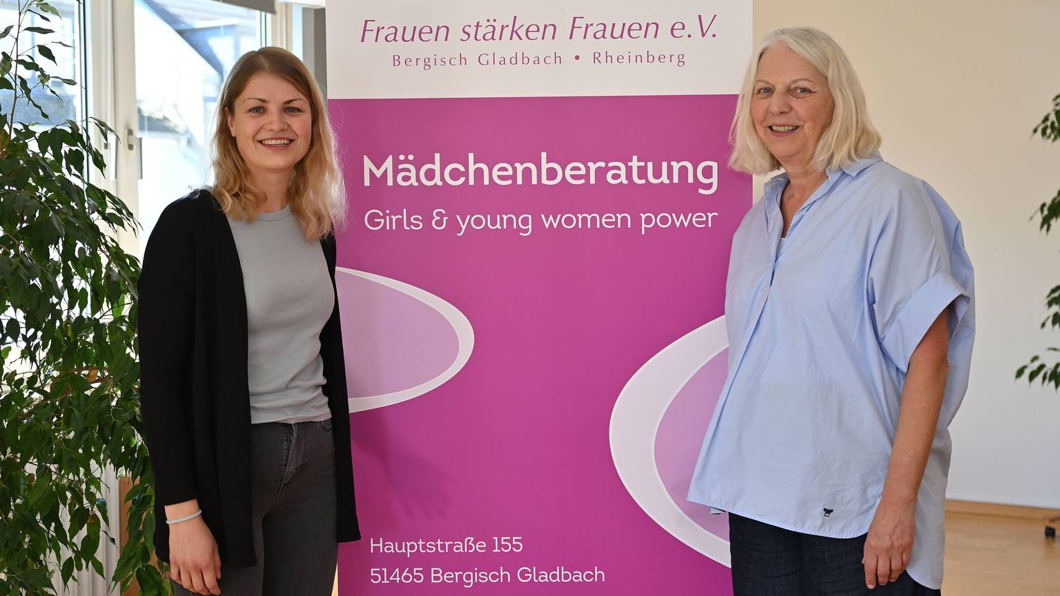 Die beiden Frauen stehen nebeneinander vor einem Plakat.