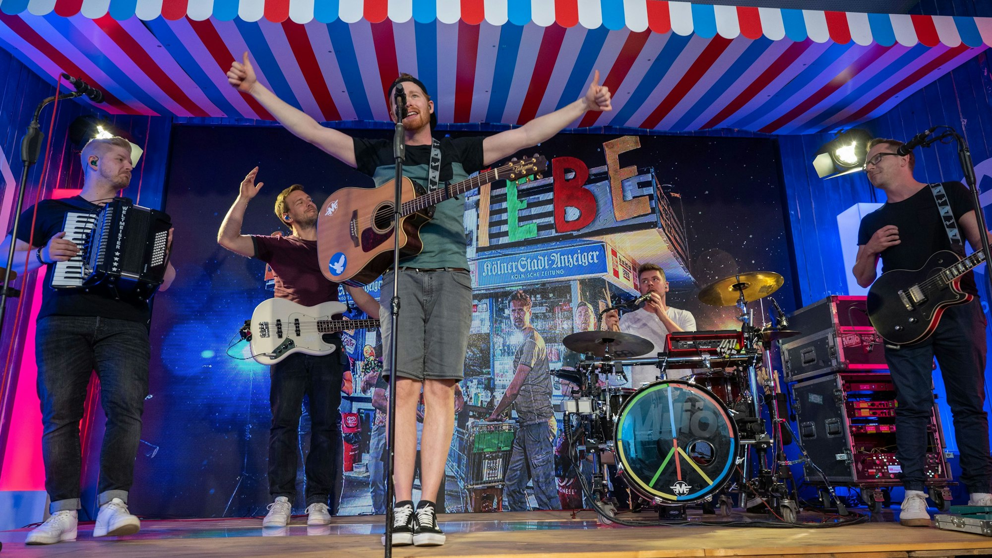 Die Band steht auf der Bühne und spielt. Der Sänge streckt die Arme aus und hält beide Daumen nach oben.
