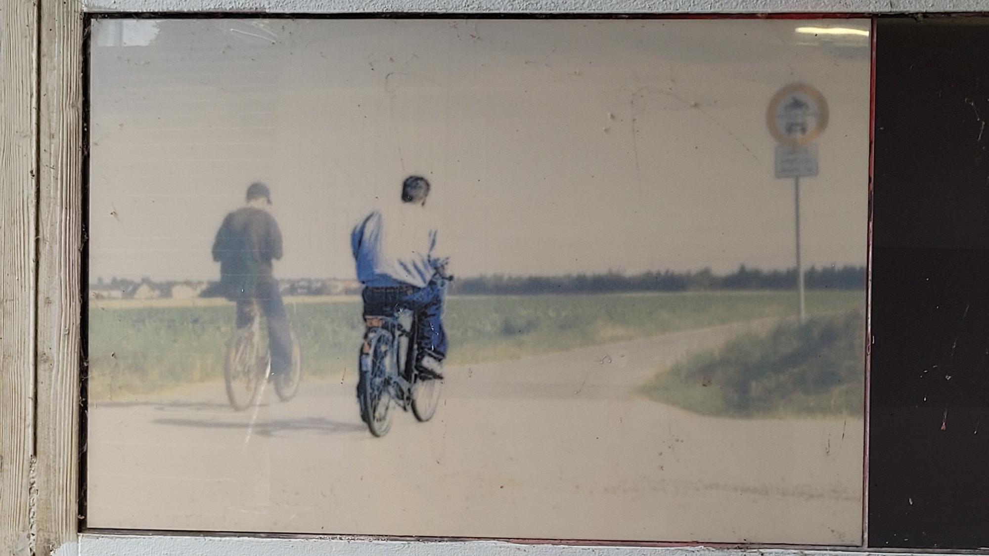 Zwei Männer auf Fahrrädern auf einem Weg zwischen Feldern.