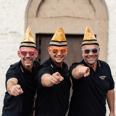 Das Bild zeigt die drei Männer vor der Flamersheimer Kirche. Sie tragen bunte Sonnenbrillen und eine Narrenkappe.