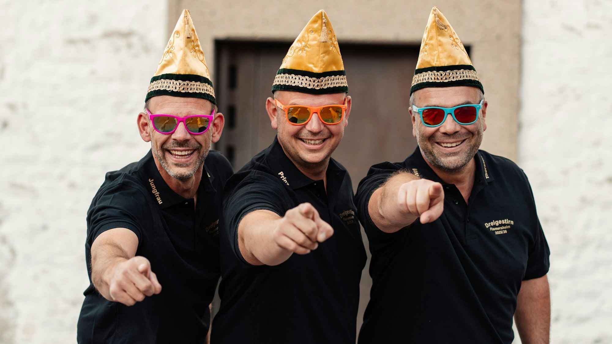 Das Bild zeigt die drei Männer vor der Flamersheimer Kirche. Sie tragen bunte Sonnenbrillen und eine Narrenkappe.