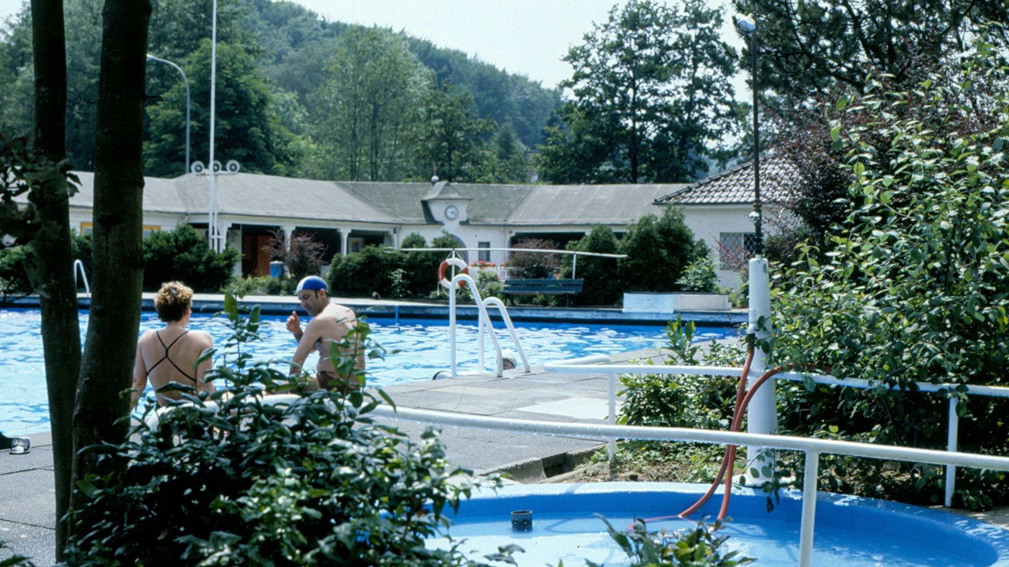 Eine alte Farbaufnahme des Freibads Herrenstrunden mit zwei Wasserbecken und Gästen.