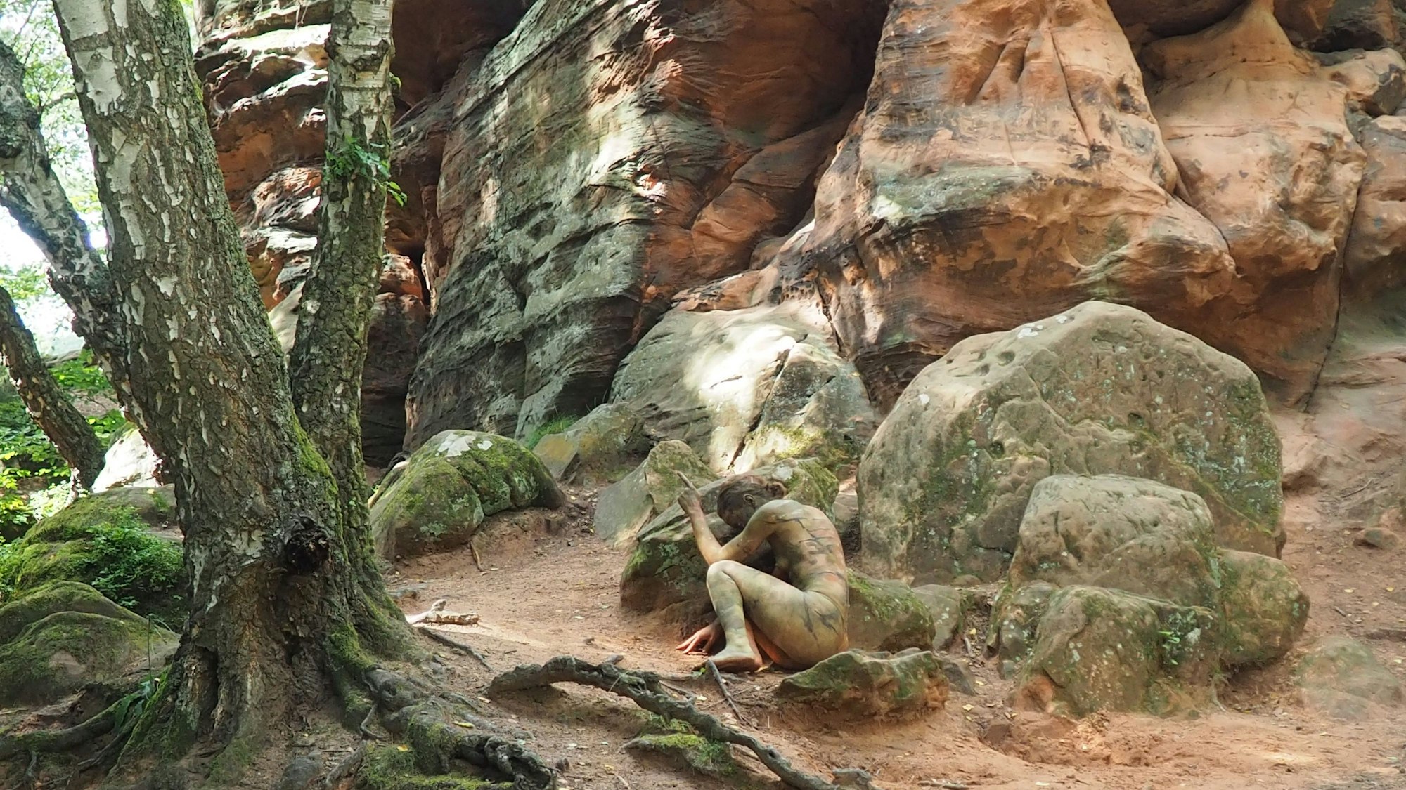 Die in Erdfarben bemalte junge Frau kauert so an einem Stein, dass sie sich in die Landschaft einfügt.