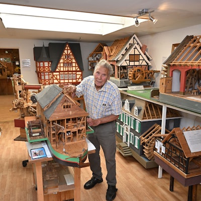 Ein alter Mann steht in einem Raum mit zahlreichen Holzmodellen von Häusern und Mühlen