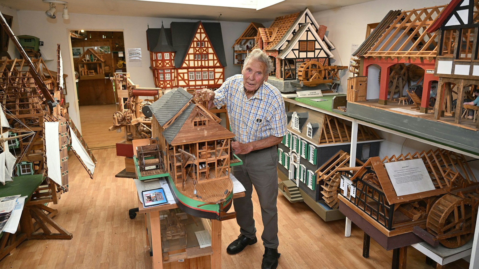 Ein alter Mann steht in einem Raum mit zahlreichen Holzmodellen von Häusern und Mühlen
