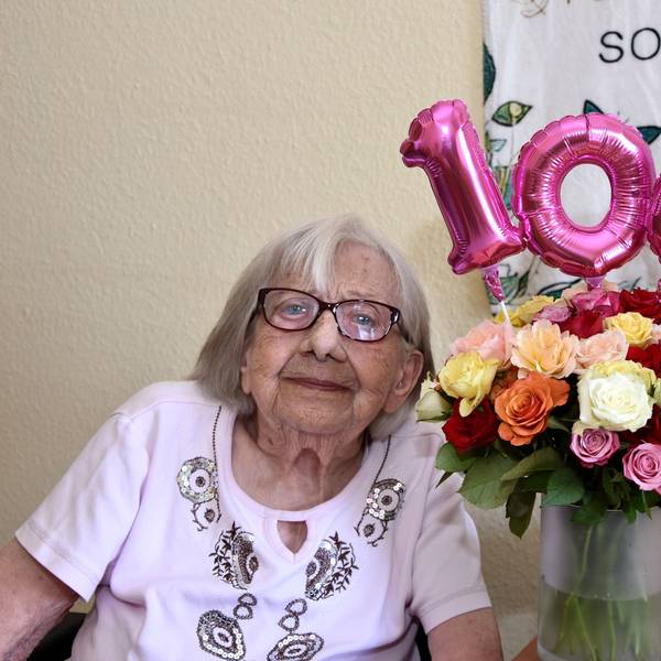 Neben der Seniorin steht eine Vase mit Blumen, darüber die Zahl 100 als Luftballon.