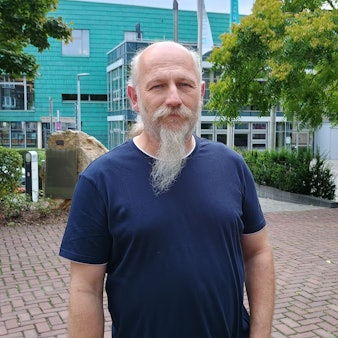 Peter Hirseler trägt ein dunkelblaues T-Shirt und hat einen Bart. Hinter ihm sieht man einige Bäume und das Medio Rhein Erft.