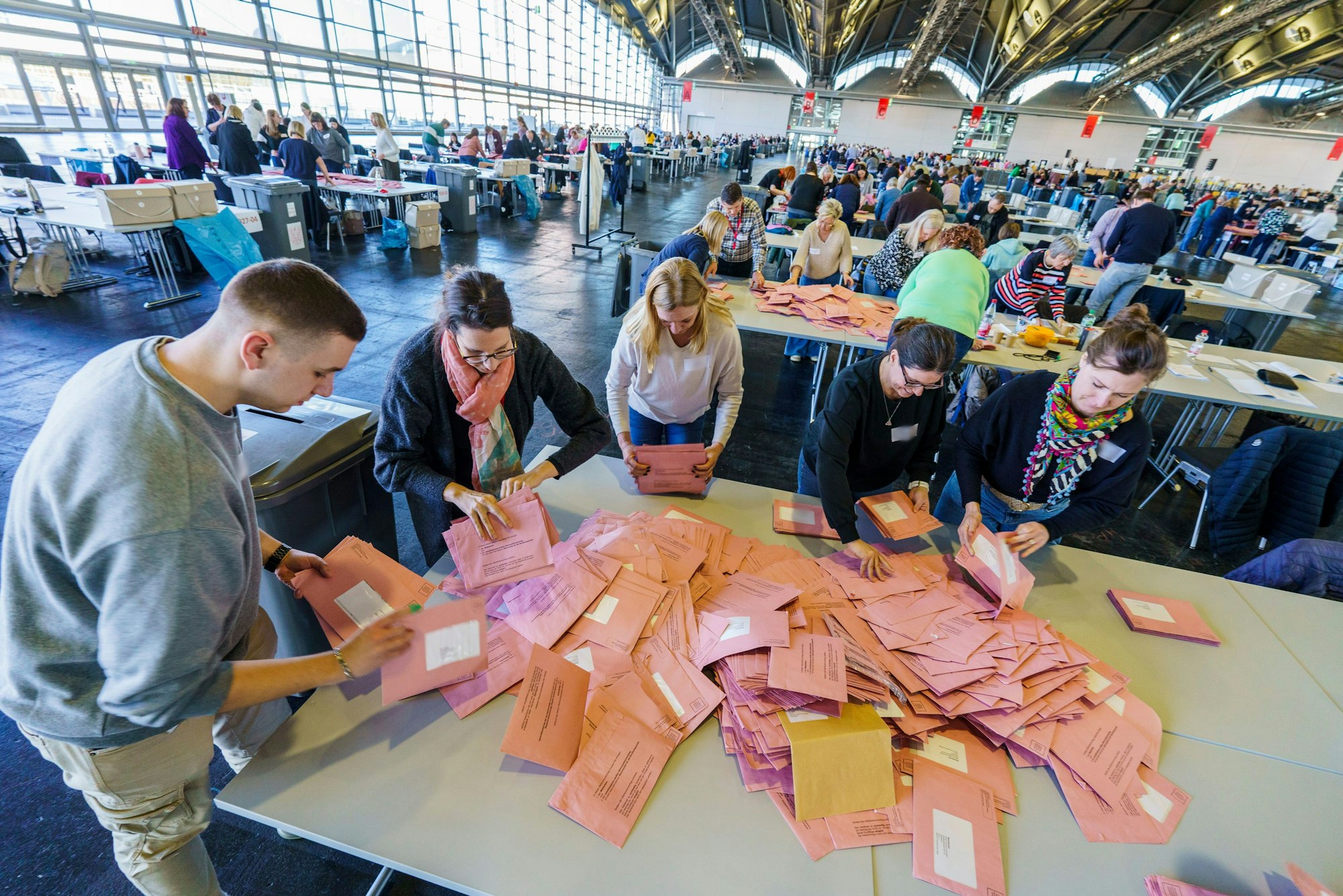 Das Bild zeigt eine Halle mit mehreren Tischen, auf den Wahlumschläge liegen. Zahlreiche Wahlhelfer sortieren die Umschläge.