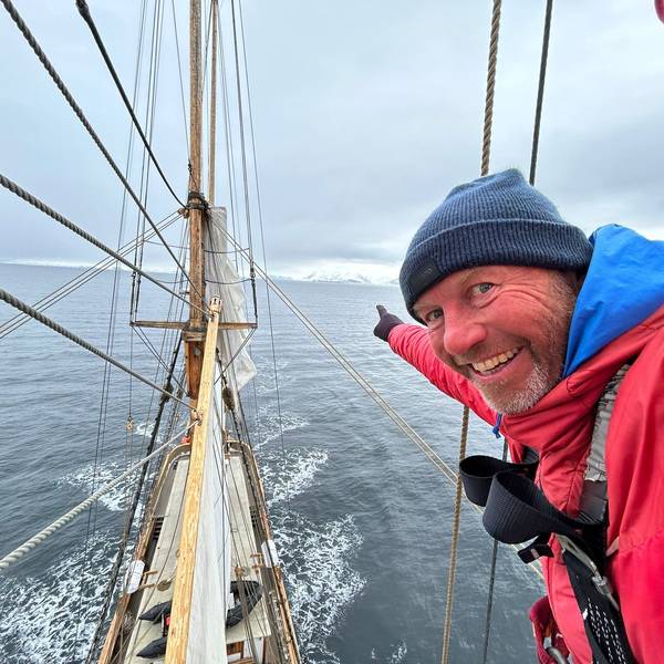 André Hauschke trägt eine rote Daunenjacke und blaue Wollmütze und deutet an Bord eines Schiffes in Richtung Horizont.