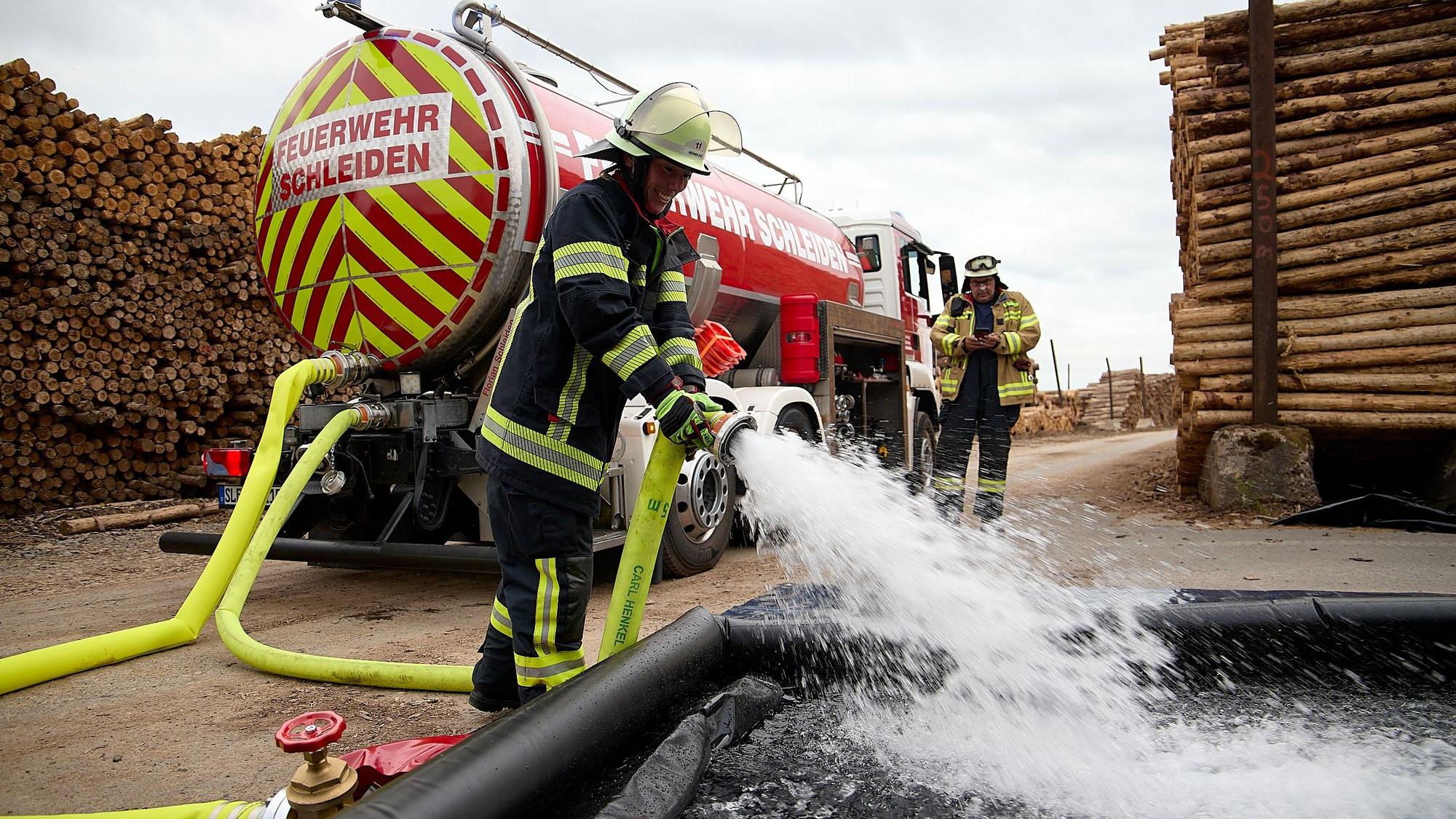 Aus einem Schlauch, den ein Feuerwehrmann hält, spritzt Wasser heraus. Im Hintergrund sind der Tankwagen und ein weiterer Feuerwehrmann zu sehen.
