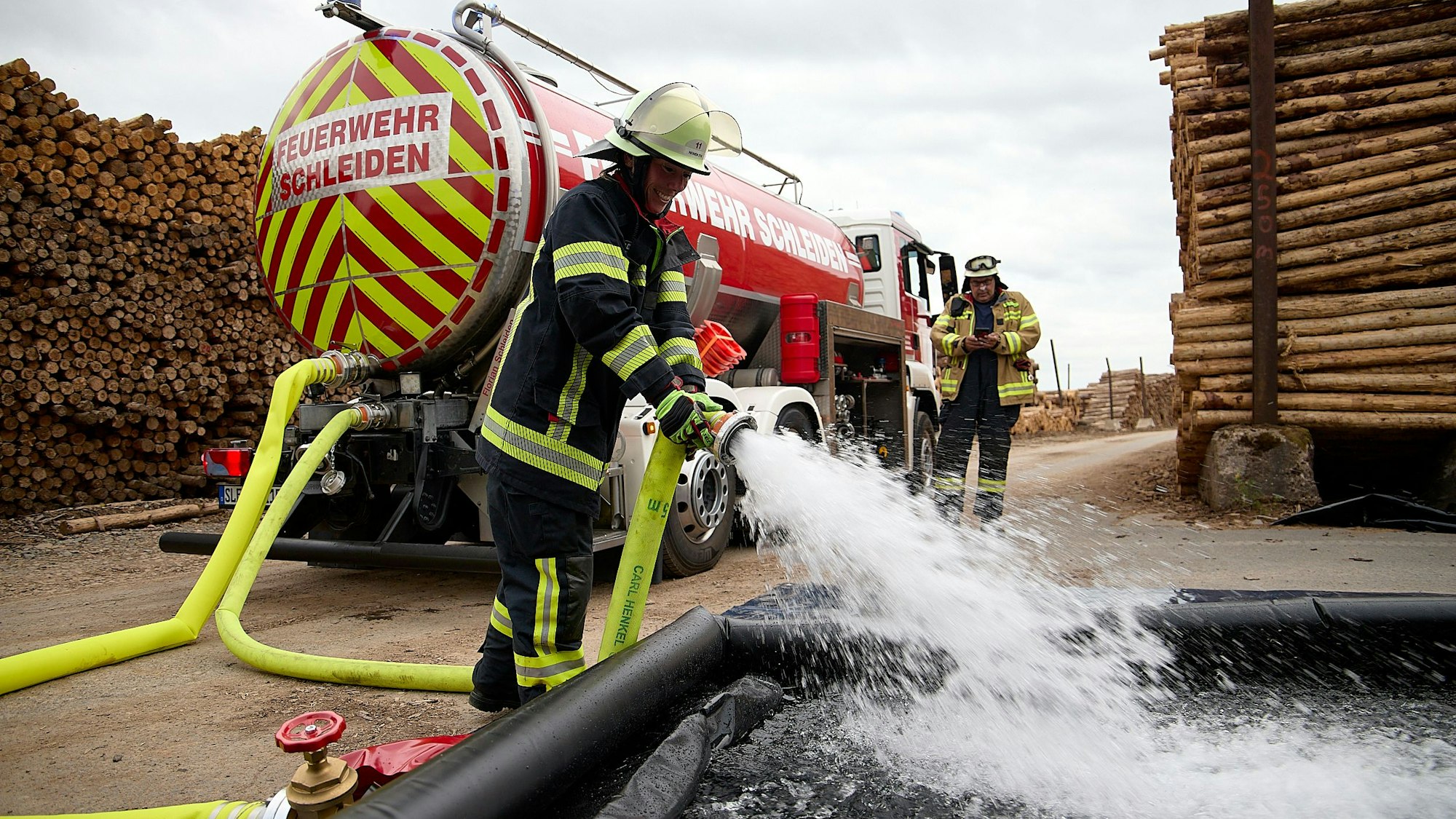 Aus einem Schlauch, den ein Feuerwehrmann hält, spritzt Wasser heraus. Im Hintergrund sind der Tankwagen und ein weiterer Feuerwehrmann zu sehen.