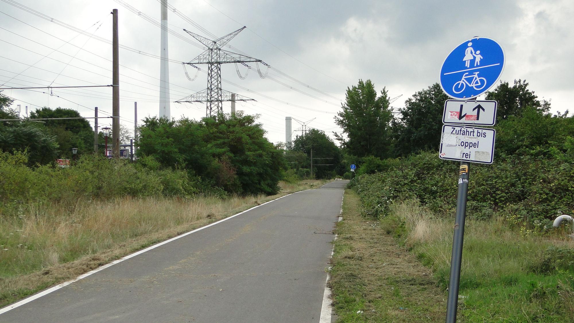An einem asphaltierten Radweg entlang einer Schienentrasse steht ein Schild, das den Weg als Rad- und Fußweg ausweist.